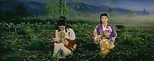 Two woman in traditional Japanese dress, in what is obviously a studio set, kneel at a memorial