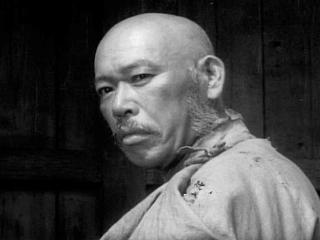 A head shot of a bald, middle-aged Japanese man in feudal-era clothes looking over his shoulder at the camera