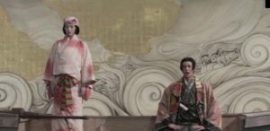 A woman and a man in traditional Japanese dress are standing and seated, respectively, in a boat against a backdrop that looks like a theatrical set