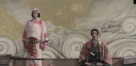 A woman and a man in traditional Japanese dress are standing and seated, respectively, in a boat against a backdrop that looks like a theatrical set