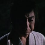 At night, a man in samurai garb with an angry-looking face in medium close-up stares at someone in front of him