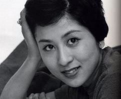 Headshot of a young Japanese woman in modern dress, her head resting upon the palm of her hand and looking into the camera with a faint smile, 