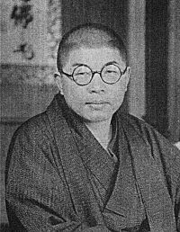 A black-and-white photo of a bespectacled Japanese man in traditional dress, looking into the camera