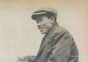 Photo of a Japanese man in a cloth cap and coat sitting in a field