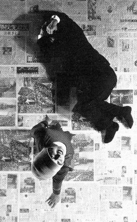 Photo from above of a man in a suit lying about a floor covered in newsprint with a woman standing over him