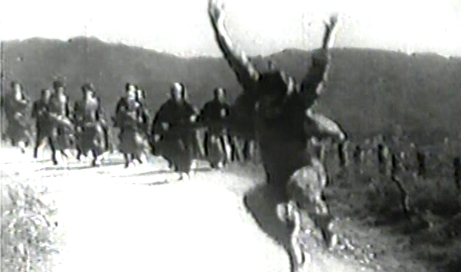 A Japanese man in a suit jumps for joy on a country road, where he is pursued by a group of men in traditional clothing