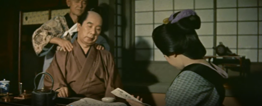 A middle-age man in 18th Century Japanese clothes is getting his shoulders massaged by a blind masseur, while a woman sits in the foreground