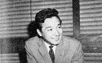 Picture of a smiling man in a jacket and tie, sitting behind a desk in from of venetian blinds