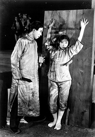 A photo of a woman bound to a wall and being threateningly addressed by a taller man, both in traditional Japanese clothing