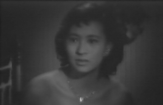 A black-and-white photo of a Japanese girl in a low-cut gown, looked anxiously off to one side