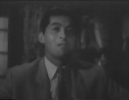 A black-and-white photo of a Japanese man in a business suit, speaking to someone off-camera
