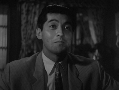 A black-and-white photo of a Japanese man in a business suit, speaking to someone off-camera