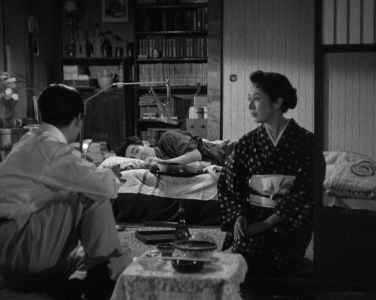 Three Japanese people in modern clothes are in a room together, in which one of the men is lying in bed in the center of the photo and another man is seated on the ground at left, as a woman at right watches the seated man uneasily