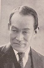 Image of a smiling middle-aged Japanese man in a suit, looking at something off-camera