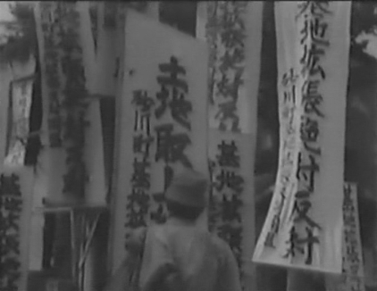A standing Japanese man, seen from behind, stares up at a group of protest banners, written in Japanese