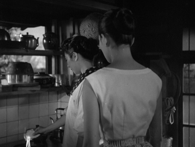 Two Japanese women - one in modern dress, the other dressed traditionally - stand in a kitchen; one burns a letter on the fire of the kitchen stove, while the other watches