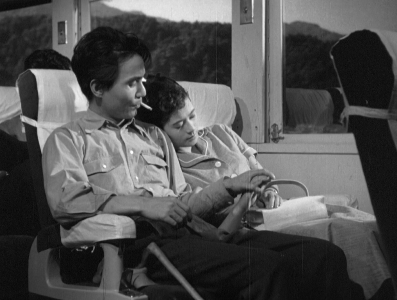 A Japanese man and woman share a seat on a train; she is sleeping and he, smoking a cigarette, removes the sleeping woman's hand from his lap