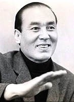 A black-and-white photo of a Japanese man in modern (ca. 1960s) dress, looking off to one side and gesturing with his hand