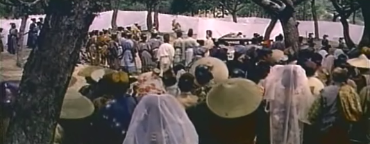 A crowd of spectators dressed as medieval Japanese stand in a field watching a man in the far distance, sleeping