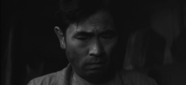 A close up of a Japanese man in working class clothes at night, looking downward in apparent great distress