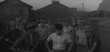 Some working class Koreans in Japan, with bicycles and wearing caps, surround a man without a cap who is looking down to the ground, lost in thought; several houses appear in the background
