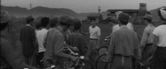 A group of men, most with bicyles, gather around their leader in a field, while a mine is visible in the background