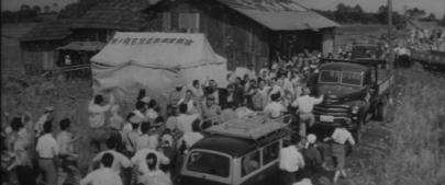 Long shot of a large jubilant crowd of Japanese people, standing in a field amid trucks, a tent and a house
