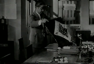 A Japanese man in a business suit in a 1930s-era office unfolds a handkerchief