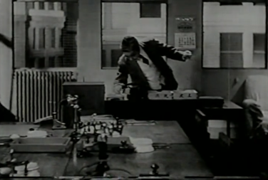 A Japanese man in a business suit in a 1930s-era office adjusts the calendar on a desk