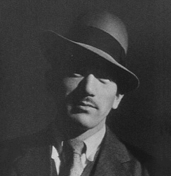 Photo of a Japanese man in his thirties, wearing a Western suit and tie and a 1930s-style hat, gazing with eyes obscured by the brim of his hat at the camera