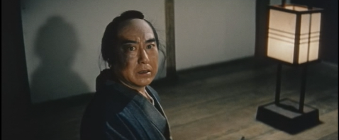 A Japanese man in feudal clothes, alone in a room and illuminated by a single lantern, looks back towards the camera, startled