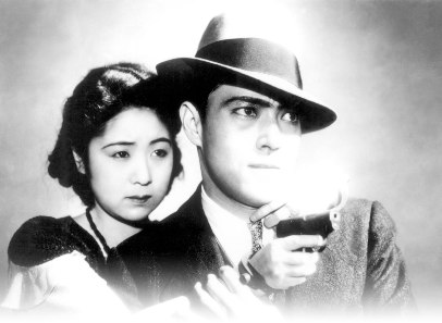 A young Japanese woman and a slightly older Japanese man standing in front of her, wearing a hat, with the latter holding a pistol, face an adversary to one side of the frame