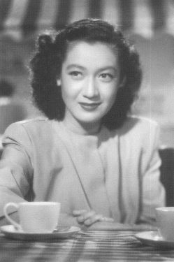 A black-and-white image of an attractive young Japanese woman in modern (1940s) clothing sitting at a table with a teacup near her elbow, looking at something slightly to one side of the camera