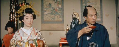 A seated woman dressed as a courtesan and a seated man dressed as a merchant in feudal Japan look disapprovingly at something off to one side, as the man holds a wine cup in his hand 