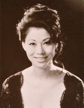 Publicity photo of a young Japanese woman smiling at the camera, wearing a piled-up hairdo and a low-cut dress