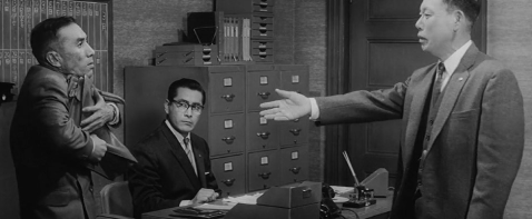A screenshot from a scene in Kurosawa's film The Bad Sleep Well, showing three Japanese businessmen in a tense confrontation