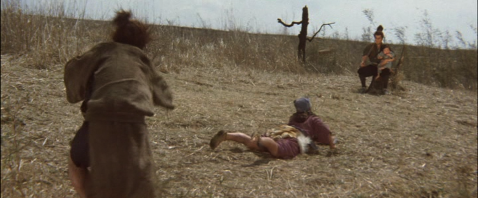 A Japanese woman in period dress, lying in a field, throws a canister at a seated man in the background holding a baby, while another man standing behind her holds a chain weapon attached to her leg