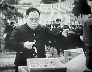 In an outdoor setting, the Japanese man in the bus uniform presents his driver's cap to another man with glasses, who looks at something inside the hat.