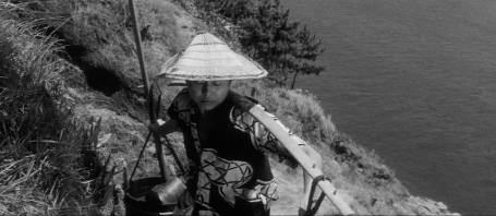 A woman in the clothes of a modern peasant, including a sedge hat, carries two buckets connected to a long pole across her shoulders up a steep hill