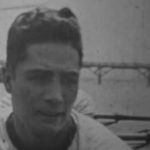 Headshot of a young Japanese man in the 1920's, setting on an unseen boat in the river, with a bridge visible behind him.