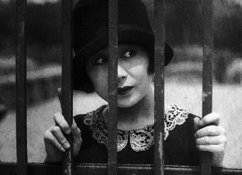 A 1920s Japanese woman in modern dress gazes at something off to the side through the bars of a gate