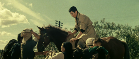 Picture of an Ainu man in modern dress on horseback against an Hokkaido landscape, giving gifts to people surrounding his horse