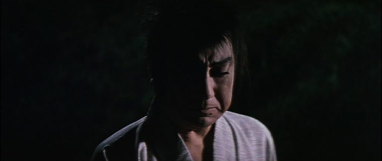 At night, a man in samurai garb with an angry-looking face in medium close-up stares at someone in front of him