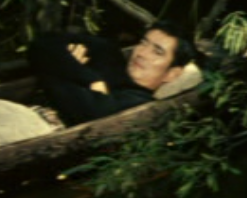 A young man lying in a canoe on the water, resting in the shade and looking unsmiling at the camera