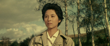 Photo of a young Japanese woman with short hair wearing a jacket, in front of a landscape of trees and hills