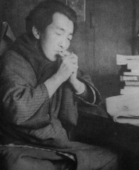 Photo of a Japanese man wearing kimono, seated in front of a desk with books and papers, and lighting a cigarette