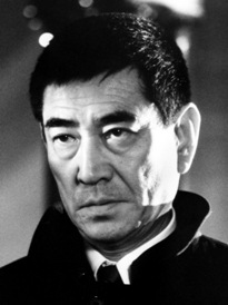 Photo of an unsmiling middle-aged Japanese man with short hair, wearing a coat with a wide collar