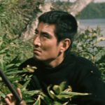 A young Japanese man in modern clothes carrying a rifle crouches behind some shrubbery and looks into the distance, with a lake in the far distance behind him