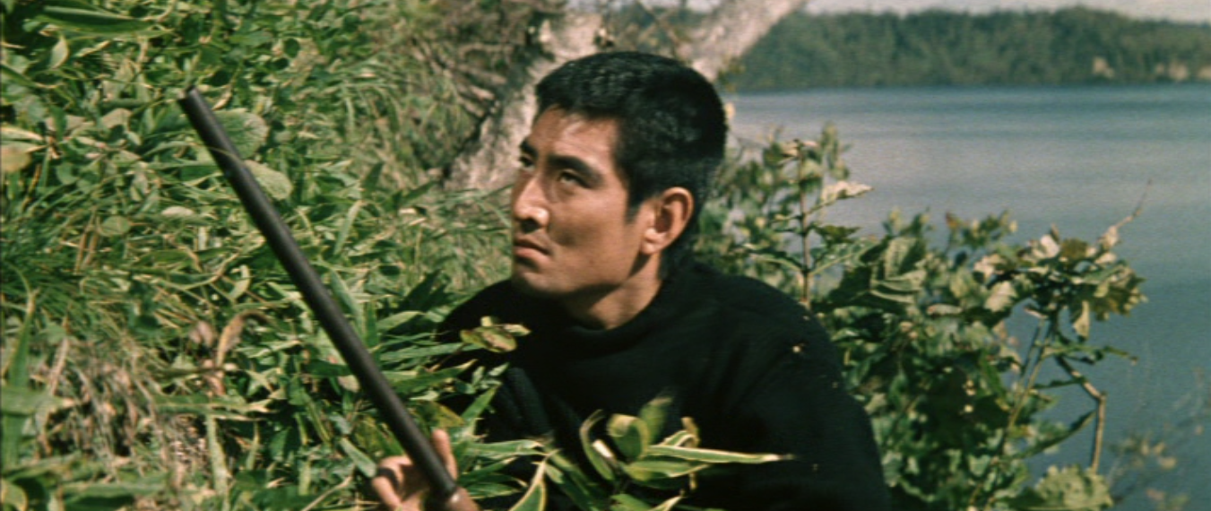 A young Japanese man in modern clothes carrying a rifle crouches behind some shrubbery and looks into the distance, with a lake in the far distance behind him