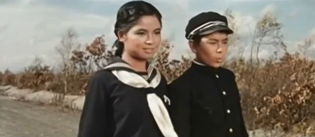 A young Japanese girl and boy, the girl slightly older, walk down a road, the girl smiling and the boy whistling